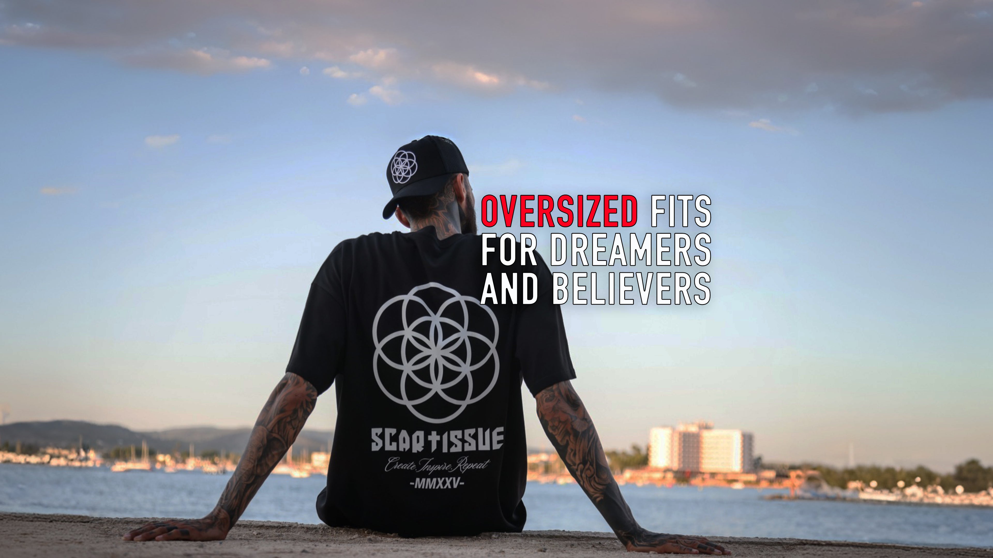 Man with tattoos wearing a black ScarTissue Core Noire Trucka hat and tee with a Seed Of Life  logo and text, sitting by a waterfront with a cityscape in the background.