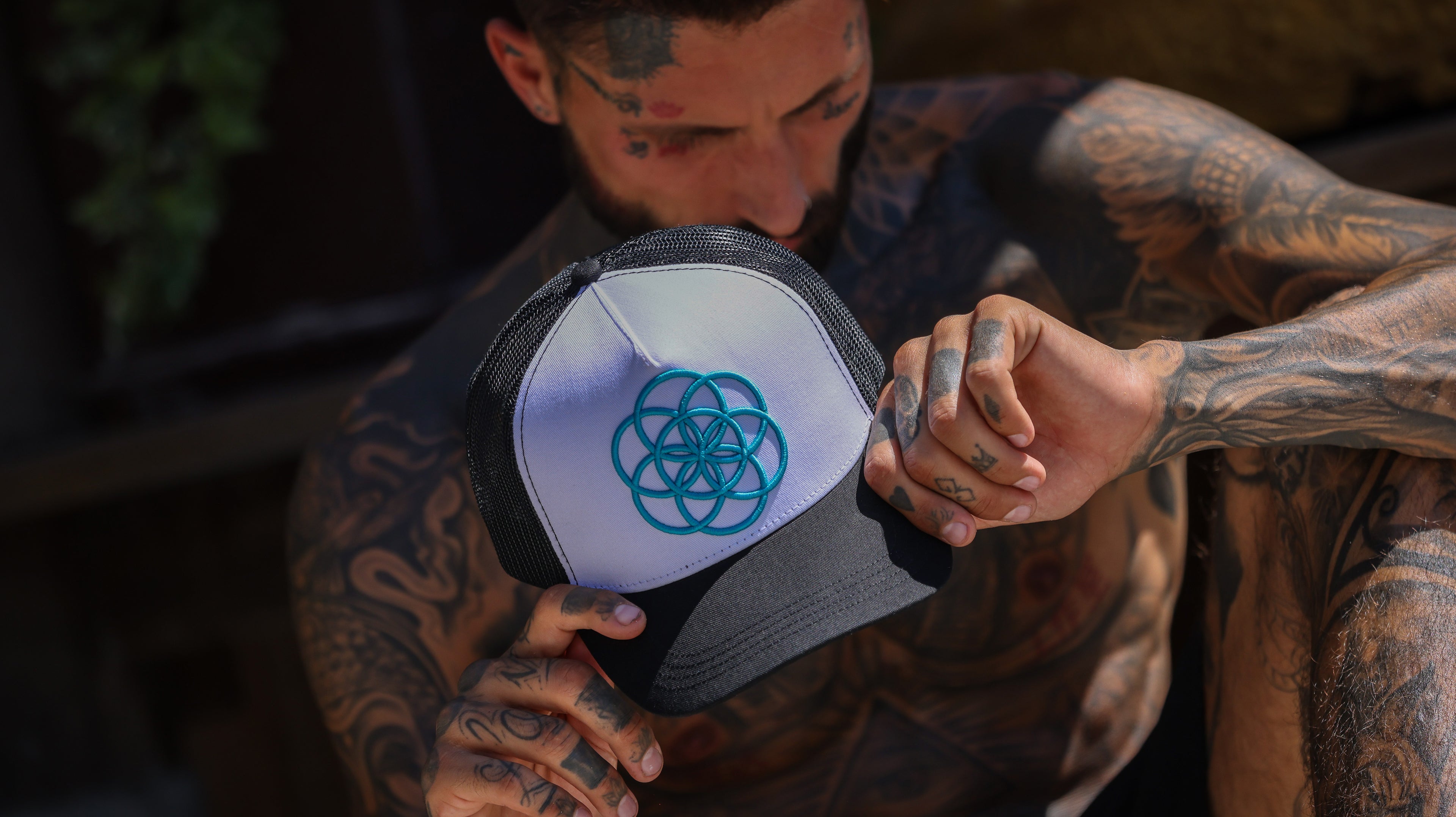 Man with tattoos holding a ScarTissue Clothing cap with a Seed Of Life logo