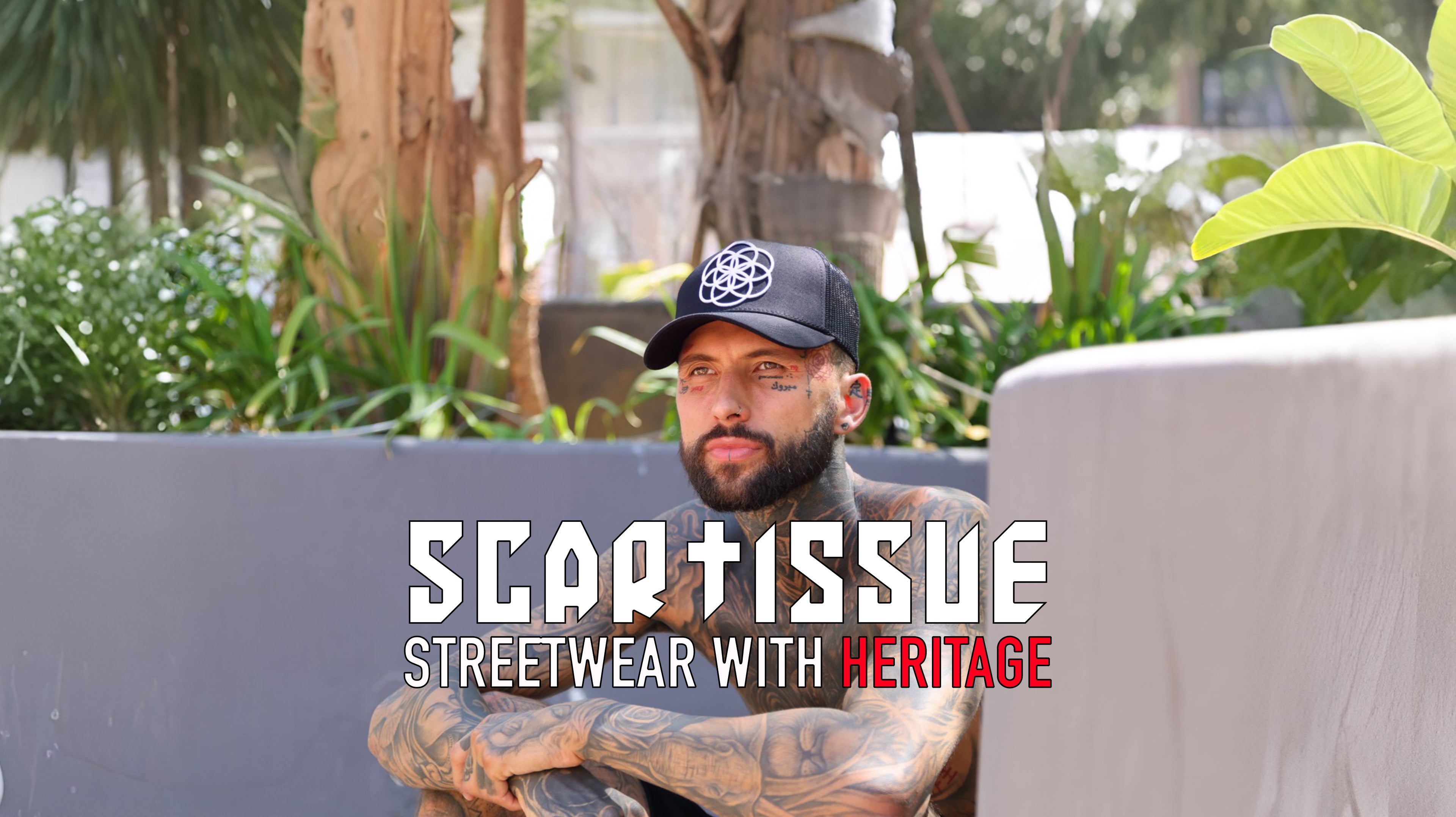 Man with tattoos sitting outdoors with black Scartissue Clothing Trucka hat streetwear branding displayed