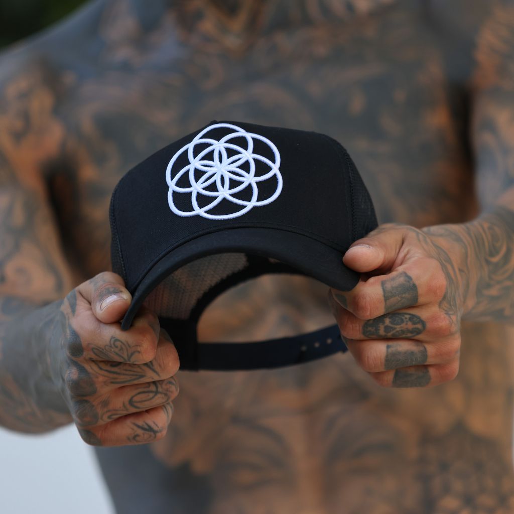 Heavily tattooed man holding a black hat with a white embroidered logo, made by scartissue clothing